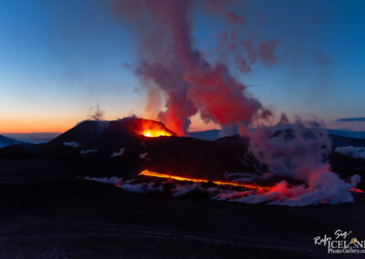 A dramatic volcanic eruption scene under a twilight sky. The image captures a volcano spewing glowing lava and thick smoke. The lava flows down the slope, illuminated in orange and red hues, contrasting against the dark landscape and the fading light of day. Billowing clouds of ash and steam rise into the sky, while distant mountains are faintly visible in the background.