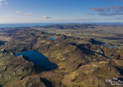 Aerial view of a rugged landscape featuring rolling hills, valleys, and two blue lakes. The terrain is a mix of brown and green hues, with patches of moss visible. In the distance, the ocean can be seen along with more hills, creating a vast, open atmosphere under a clear blue sky with a few scattered clouds.