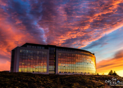 A modern building with large glass windows reflects a vibrant sunset. The sky displays shades of pink, orange, and blue, with dramatic clouds adding depth to the scene. The structure has a rounded roof and is set against a backdrop of greenery.