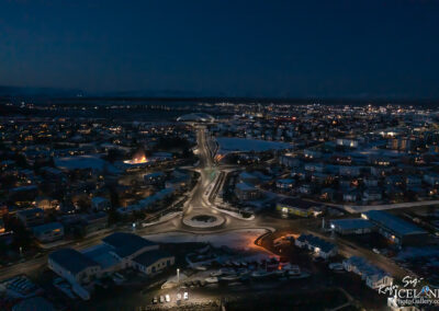 A nighttime aerial view of a town illuminated by scattered lights. The scene features residential buildings, a large roundabout at the center with several vehicles, and a snowy landscape. In the background, the illuminated structures of the town stretch toward the horizon, while faint mountains are visible under a dark blue sky. A few boats are docked near the bottom of the image.