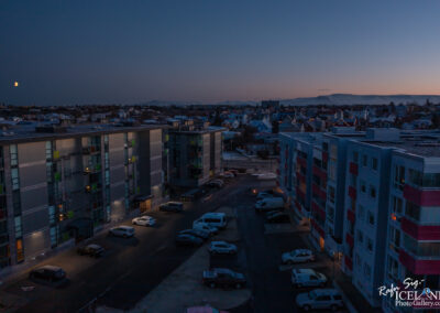 A twilight scene featuring an urban neighborhood. In the foreground, several modern apartment buildings with illuminated windows are visible, surrounded by a parking lot filled with various vehicles. In the background, a panoramic view of the city and houses can be seen, with distant mountains looming under a darkening sky. A crescent moon is faintly visible in the upper left corner, adding a serene touch to the evening atmosphere.