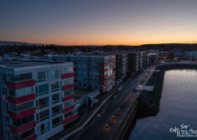 A twilight view of modern apartment buildings along a waterfront. The structures feature large windows and balconies with red accents. A pathway lined with soft lights runs beside the water, which reflects the fading orange and blue hues of the sunset. In the background, hills and additional buildings are visible under a gradually darkening sky.