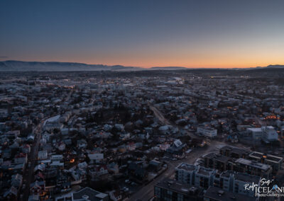 A panoramic view of a city at dusk, showcasing a mix of residential and commercial buildings. The houses are clustered closely together, with some illuminated by streetlights and windows. In the background, a mountain range is visible under a twilight sky with soft gradients of blue and orange. The cityscape features winding roads, hinting at the urban layout, and the soft glow from lights adds a serene atmosphere to the scene.