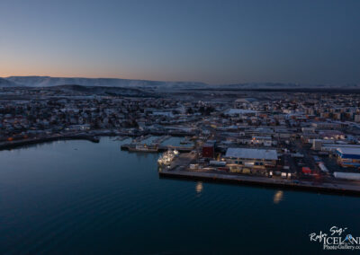Aerial view of a coastal town at dusk, featuring a harbor with several boats docked. The waterfront is lined with a mix of residential and commercial buildings, illuminated by soft lights. In the background, snow-capped mountains are visible under a twilight sky, creating a serene atmosphere. The water reflects the fading light, adding to the tranquil scene.