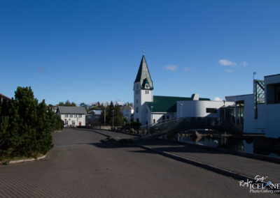 A tranquil urban scene featuring a wide, clear blue sky above a paved pathway lined with low greenery and modern architecture. Prominently displayed is a white church with a tall green spire and clock, alongside a sleek modern building. A small pedestrian bridge arches over a still body of water, enhancing the serene atmosphere of the area.