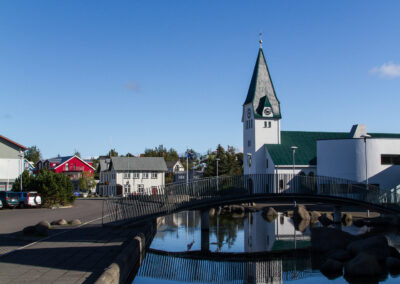 A calm, sunny day in a small town featuring a white church with a green roof and a prominent steeple. The church is reflected in a clear body of water in the foreground, while a small modern bridge crosses the water. Surrounding buildings include colorful houses and parked cars, with trees in the background under a bright blue sky.