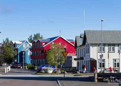 A vibrant street scene featuring colorful buildings in various styles, including a red and blue house, with a clear blue sky above. The foreground shows a roundabout with a yield sign, decorative planters, and a few people walking, including a woman in a red coat and a child. Parked cars are visible, and greenery is seen around the structures, adding to the charm of the area.