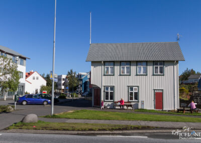 A quaint, two-story white house with a corrugated metal roof is set against a clear blue sky. The building features several windows with green frames and a red door. In front of the house, two women and a child are seated on benches, enjoying the outdoors. The area around the house has green grass and a few small trees. To the left, additional buildings in various colors can be seen, along with a parked blue car and street signs.