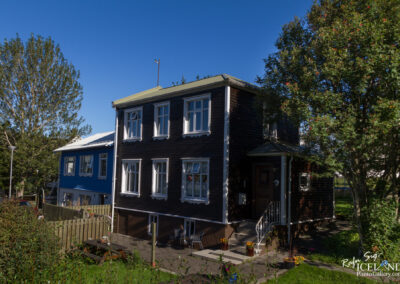 A two-story house with a brown wooden exterior and white window frames sits beside a blue house. The house features a small staircase leading to the front door, with two chairs placed near the entrance. In the foreground, there is a wooden fence and some greenery with flowers, while a tall tree with green leaves stands nearby against a bright blue sky.
