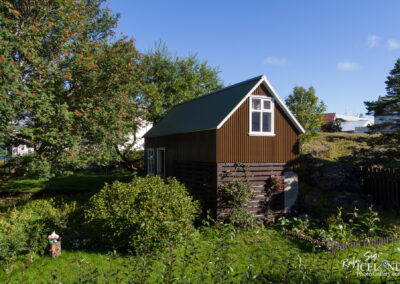 A small, quaint brown house with a gabled roof is surrounded by lush green foliage and trees, including one displaying bright red berries. The house features white window frames and is partially obscured by shrubs and plants in the garden. In the background, additional houses and a clear blue sky are visible, indicating a serene outdoor setting.