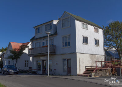 A three-story building with a mixture of white and green siding, featuring multiple windows and a balcony. A small wooden deck is visible at the front, along with outdoor seating. To the left, there’s a smaller white house with a red roof and a tree in front. Cars are parked on the street in front of the building under a clear blue sky.