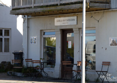 A quaint building with a sign reading "Gamla Matarbúðin" is featured, indicating a food outlet. The exterior has a light-colored wall showing signs of wear. There are several empty wooden chairs arranged outside, with a small table positioned among them. To the left, a trash bin is placed next to the building. The window displays some greenery, reflecting the surroundings. It is a sunny day, casting shadows on the ground.