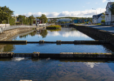A tranquil waterway flows through a scenic area, flanked by pathways and greenery. Small stone structures create gentle cascades within the water, while reflections of the blue sky and fluffy clouds can be seen on the surface. Beside the water, buildings line the way, with trees and shrubs adding natural beauty to the urban landscape.