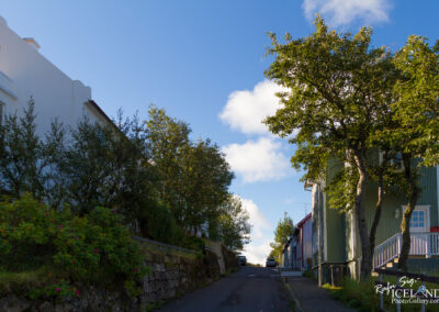 A narrow street lined with colorful houses, some featuring green and red facades, and flanked by bushes and trees. The sky is clear with a few fluffy clouds, and a car is parked further down the street, creating a picturesque, serene atmosphere.