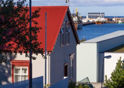 A view of a coastal town with a red-roofed house in the foreground, featuring a white picket fence and greenery. In the background, a harbor is visible with several boats docked, and the horizon shows a clear blue sky with a few clouds. Street lamps are positioned along the path leading towards the harbor.