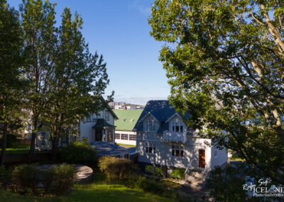 A picturesque residential scene featuring two distinct houses nestled among lush green trees. The house on the left has a traditional architectural style with a sloped roof and decorative elements, while the house on the right has a more modern look with a light-colored exterior and a welcoming porch. The background reveals a clear blue sky and distant urban buildings, suggesting a vibrant neighborhood atmosphere. A circular wooden structure and well-maintained landscaping add to the charm of the setting.