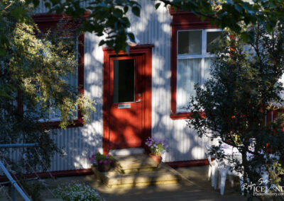A close-up view of a house featuring a red door with a glass panel and matching red window frames. The house has a corrugated metal exterior and is surrounded by greenery, including a bush with small leaves and colorful flowers in pots on the steps. A wooden deck with a table and chairs is partially visible to the right. Soft sunlight filters through the leaves, casting shadows on the exterior.