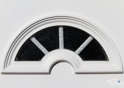 A close-up of a decorative arch-shaped window featuring a black background with shimmering silver accents, framed by a white border. The design includes vertical and horizontal lines creating a geometric pattern within the arch.