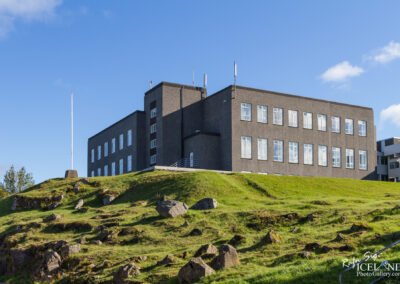 A modern, two-story building with a gray exterior and many windows, situated on a grassy hill. There are several rocks scattered on the slope leading up to the building, and a flagpole stands to the left. The sky is clear with a few clouds visible.