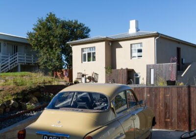 A vintage yellow car is parked in a driveway, facing away from the camera. In the background, a beige house with multiple windows and a chimney is visible, along with a wooden fence and a tree. The sky is clear and blue, indicating a sunny day. A bicycle is leaning against the fence, and there are some shrubs and rocks in the foreground.