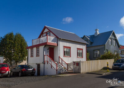 A charming two-story white house with red accents, featuring a front porch and a staircase leading to the entrance. Behind the house is a green building and a lush tree. Two parked cars, one red and one gray, are visible in the foreground on a gravel surface. The sky is clear with a few wispy clouds.