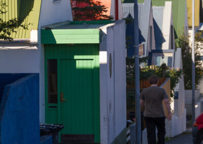 A narrow street lined with colorful, pitched-roof houses in various shades of green, orange, and blue. A person walks away from the camera along the sidewalk, passing by a green door on a white structure, with trash bins nearby. The scene captures a sunny day with clear skies and a hint of the ocean in the background.