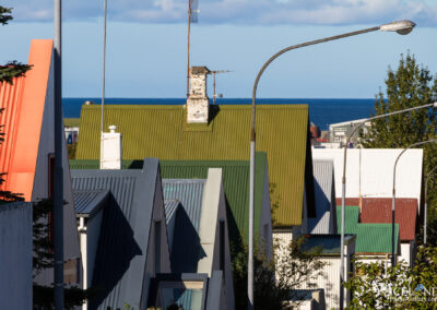A view of a row of rooftops in various colors, including orange, green, grey, and white, against a blue sea in the background. The scene is completed with streetlights and some greenery in the foreground. The sky is partly cloudy.