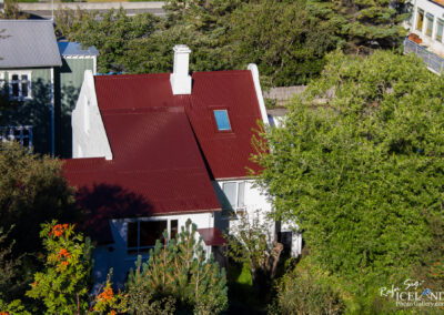 An aerial view of a white house with a red corrugated metal roof, featuring a blue window near the peak. Surrounding the house are lush green trees and shrubs, with some colorful flowers visible. In the background, there are structures with gray and green facades, indicating a residential area. The scene suggests a bright, sunny day.