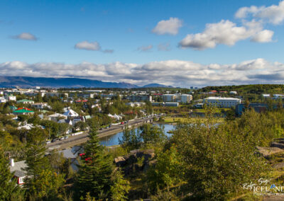 A scenic view of a town with various buildings, including houses with colorful roofs and modern structures, set against a backdrop of mountains and a partly cloudy sky. Lush greenery and trees frame the foreground, while a river runs along the edge of the town, with a road beside it. The atmosphere is bright and serene, typical of a clear day in a rural or suburban setting.