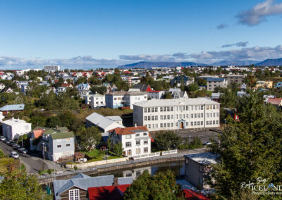 A scenic view of a residential area in Reykjavik, Iceland, featuring a mix of colorful houses with pitched roofs, surrounded by lush greenery. In the foreground, a large white building stands prominently, complemented by nearby trees and a winding street. The background showcases more houses and hills under a clear blue sky, with a few clouds scattered above.