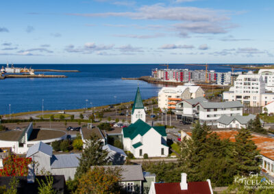 A panoramic view of a coastal town featuring a mix of modern and traditional architecture. In the foreground, a white church with a green steeple stands prominent. There are several residential buildings, some with colorful facades, along the waterfront. The calm blue sea stretches out into the horizon, with boats and a harbor visible. A clear sky with scattered clouds adds to the serene atmosphere.