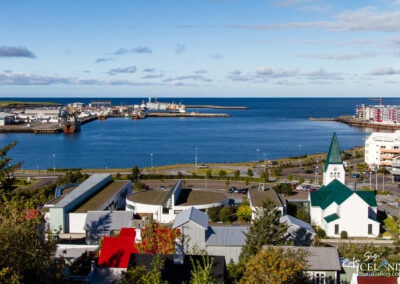 A panoramic view of a coastal town featuring a mix of modern and traditional architecture. In the foreground, a white church with a green steeple stands prominent. There are several residential buildings, some with colorful facades, along the waterfront. The calm blue sea stretches out into the horizon, with boats and a harbor visible. A clear sky with scattered clouds adds to the serene atmosphere.
