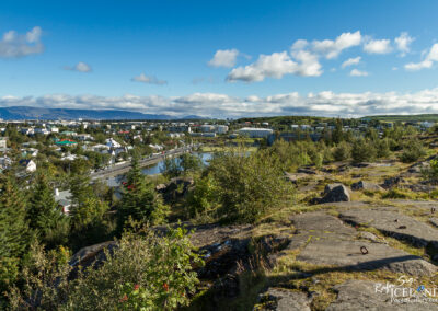 A panoramic view of a town with colorful buildings and greenery, set against a backdrop of rolling hills and a blue sky with scattered clouds. In the foreground, rocky terrain with patches of grass and small trees leads to a small river that runs through the town, which features a mix of modern and traditional architecture.