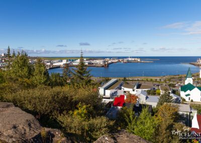 A panoramic view of a coastal town, showcasing a harbor filled with boats and industrial buildings. In the foreground, various rooftops in shades of red, white, and green are visible amidst lush greenery. A white church with a green steeple stands out among the buildings. The backdrop features a clear blue sky with a few clouds, and the calm sea extends to the horizon.
