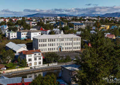 A panoramic view of a small town featuring a mix of colorful houses and buildings. In the foreground, there is a large, multi-story light-colored building with numerous windows and a central entrance. Surrounding it are various residential structures with sloped roofs in shades of red, blue, and white. Lush greenery is visible, along with a body of water reflecting the scene. In the background, distant mountains and a partly cloudy sky complete the picturesque setting.