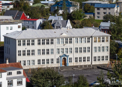 A large, two-story building with a white exterior and numerous windows, situated in an urban area. The entrance features a blue door and the building has a flat roof. Surrounding the structure are various other buildings with colorful roofs, trees, and a playground area. The setting appears to be a residential neighborhood with a clear blue sky overhead.