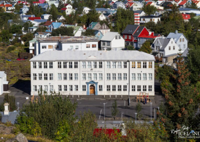 A large, three-story building with a flat roof, featuring multiple windows and a central entrance adorned with a blue door. The façade is mainly white, with some wear visible. Surrounding the building are smaller residential structures with various colored roofs, including red and green, amidst trees and a parking area. The scene is set in a sunny environment, showcasing a mixture of architectural styles typical of a town setting.