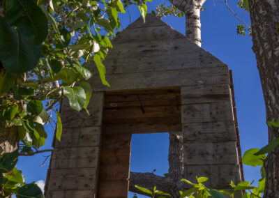 A wooden structure resembling a house, framed by vibrant green leaves and tree trunks. The sky is clear and blue in the background, creating a serene, natural setting. The wooden structure has an open doorway and a triangular roof, partially obscured by leaves.