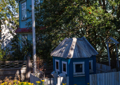 A small blue playhouse with a wooden roof is situated in a garden filled with vibrant yellow flowers. Lush green foliage surrounds the playhouse and there is a wooden fence partially visible. In the background, a pastel-colored building with a red roof and white windows can be seen, under a clear blue sky.