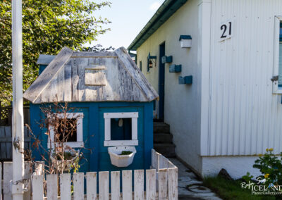 A small, colorful playhouse with a blue exterior and a slanted wooden roof stands next to a white building. The playhouse features two square windows with white frames, and a small planter box attached below one window. The area is surrounded by a white picket fence, and there are steps leading to a door on the white building, which is marked with the number 21. Green foliage and flowers are visible in the foreground, contrasting with the structures.