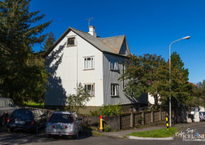 A two-story, light gray house with a corrugated steel exterior and a weathered roof sits on a corner lot. The house features multiple windows, some with broken panes, and is partially shaded by nearby trees. In the foreground, several parked cars are visible, along with a red fire hydrant and a decorative stone fence surrounding the property. The sky is clear and blue, indicating a sunny day.