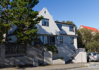 A quaint, two-story house with a white exterior and a distinctive triangular roof is surrounded by lush green trees. The house features multiple windows, some with decorative curtains visible. There is a small garden area with flowering plants near the entrance, and a set of stairs leading up to a porch. A concrete wall with a decorative pattern encloses part of the property, and a parked silver car is visible nearby on the street. The sky is clear and bright, indicating a sunny day.