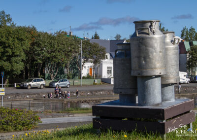 A cluster of metallic milk cans sits on a wooden platform in the foreground, with grassy areas and blooming flowers in front. In the background, there are several cars parked along a path, and groups of people walking beside a body of water. A mix of trees and buildings, including a white structure with an orange door, can be seen in the backdrop under a clear blue sky.