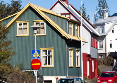 A picturesque street scene featuring a green and yellow house on the left, with multiple windows adorned by plants. Next to it, a red house has a white roof and visible satellite dishes. A signpost displaying a bicycle symbol and a stop sign is in the foreground, while various cars, including a silver one and a red one, are parked along the street. The backdrop includes clear blue skies and some trees.