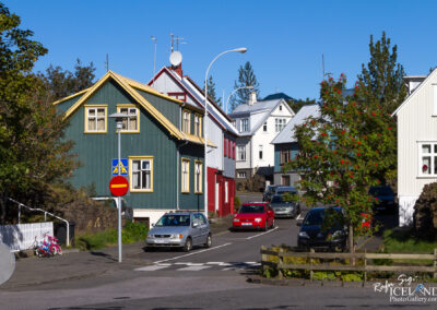A picturesque street scene featuring colorful houses with varying architectural styles. A prominent green house with yellow accents is in the foreground, along with a red house partially visible. Several cars are parked along the street, and a tree with red berries adds a touch of nature to the urban setting. Clear blue skies and bright sunlight enhance the vibrant colors of the houses and surroundings. Traffic signs are present, indicating a stop and pedestrian crossing.