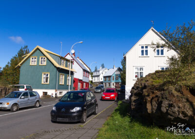 A picturesque street scene featuring a mix of colorful houses in shades of green, red, and white. The homes are set against a clear blue sky, showcasing typical Scandinavian architecture. Several parked cars line the street, including a silver hatchback and a black vehicle. In the foreground, there is a large rocky formation covered with grass. A person can be seen walking along the sidewalk, adding a sense of life to the scene.