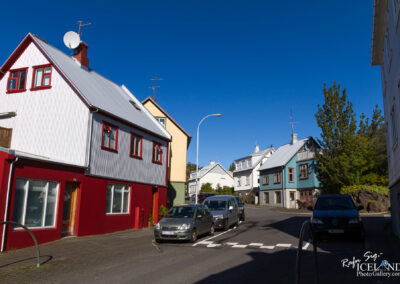 A charming street scene featuring colorful houses with metal roofs in a residential area. The building on the left is painted red with white accents, and next to it is a yellow house. The street is lined with parked cars, and the clear blue sky above adds to the picturesque atmosphere. Lush greenery is visible near the buildings, providing a vibrant contrast to the architecture.