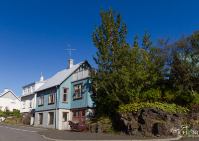 A scenic view of a residential street featuring a charming house with a blue and white exterior. The house has multiple windows and a sloped metal roof. In front of the house, there is a visible old cart, and a rock formation is visible on the right side, adorned with greenery. The backdrop is a clear blue sky, enhancing the bright and inviting atmosphere of the scene.