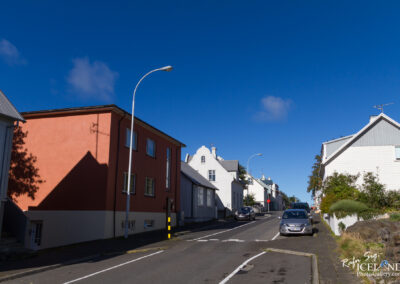 A street scene featuring a residential neighborhood with colorful houses. On the left, a bright orange building is contrasted by a white house with a gray roof. The street is lined with parked cars and streetlights under a clear blue sky, with a few scattered clouds. The road slopes slightly upward, leading into the distance where more residential buildings are visible.
