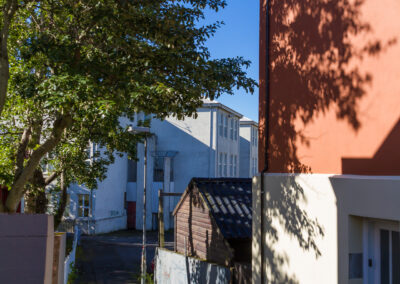 A narrow alleyway is shown, flanked by a tall brown wall on the right and a white building on the left. A large leafy tree stands on the left side, casting shadows on the ground. In the background, there are more buildings with windows facing the alley, and a small wooden shed is visible near the end of the path. The sky is bright blue with a few clouds scattered throughout.